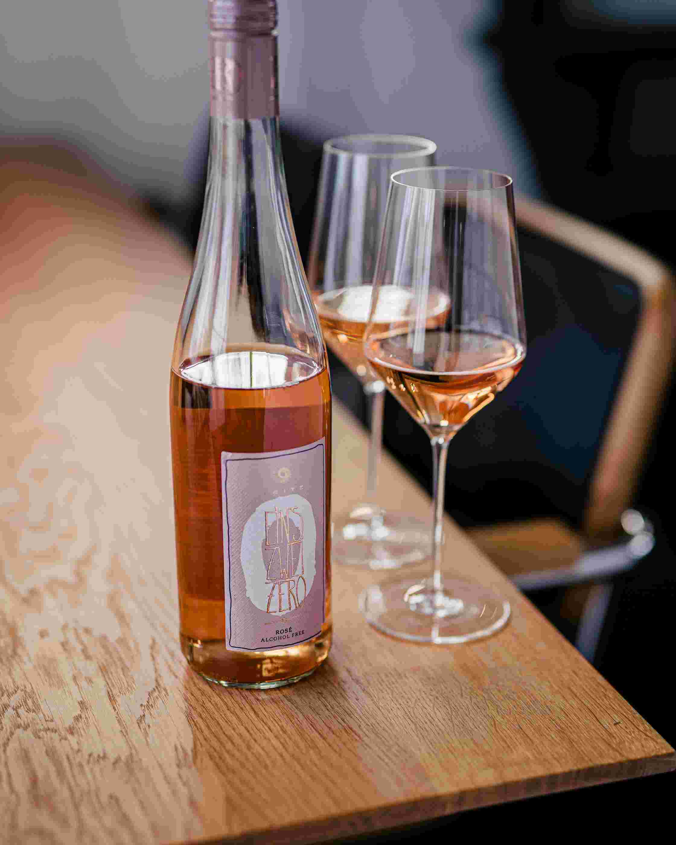 Sfeerfoto Leitz Eins-Zwei-Zero Rosé. De fles staat op een houten tafel. Naast de fles staan twee gevulde wijnglazen. 
