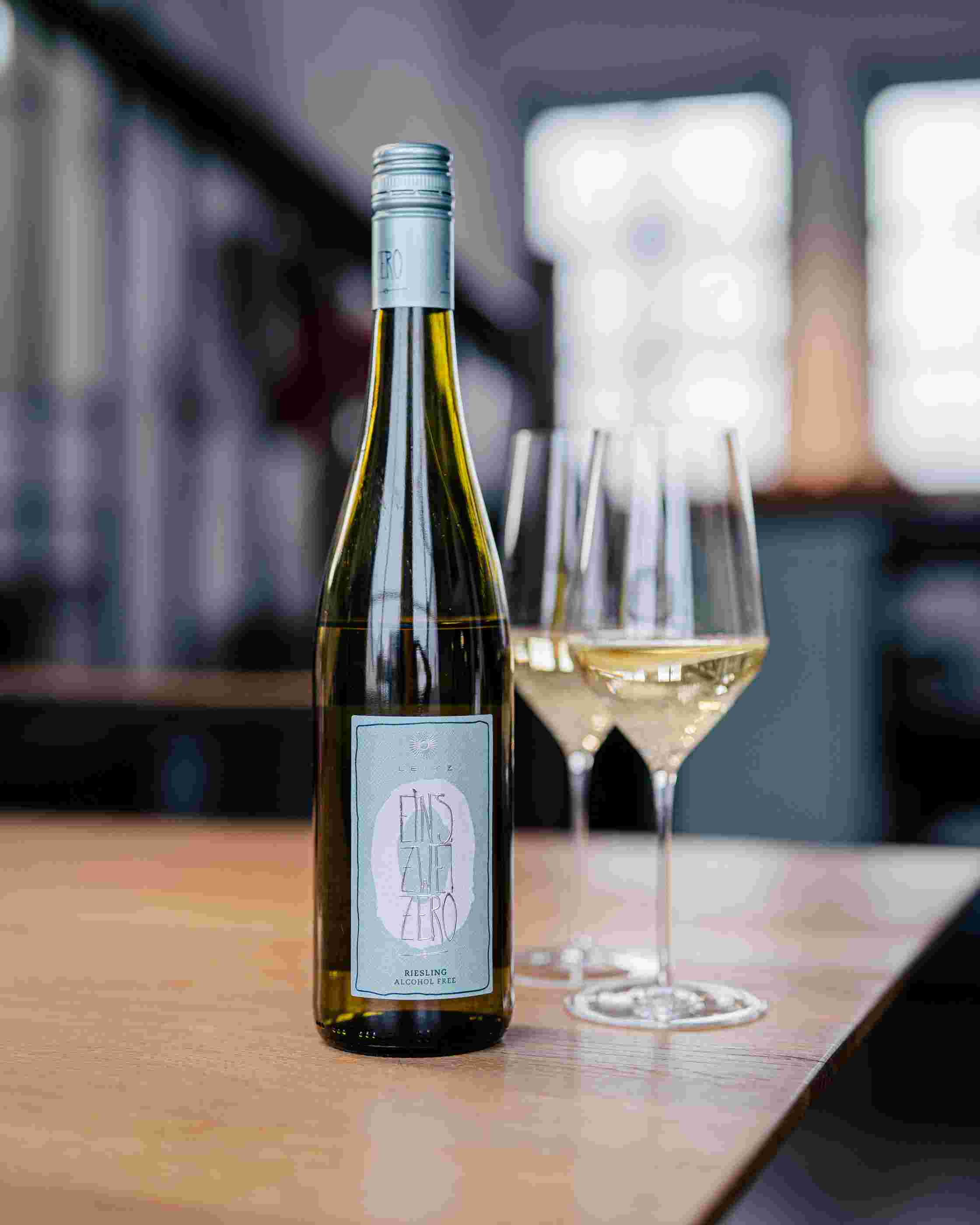 Sfeerfoto van een flesLeitz Eins-Zwei-Zero Riesling, rechts staan twee ingevulde wijnglazen op een lichtere, houten tafel.
