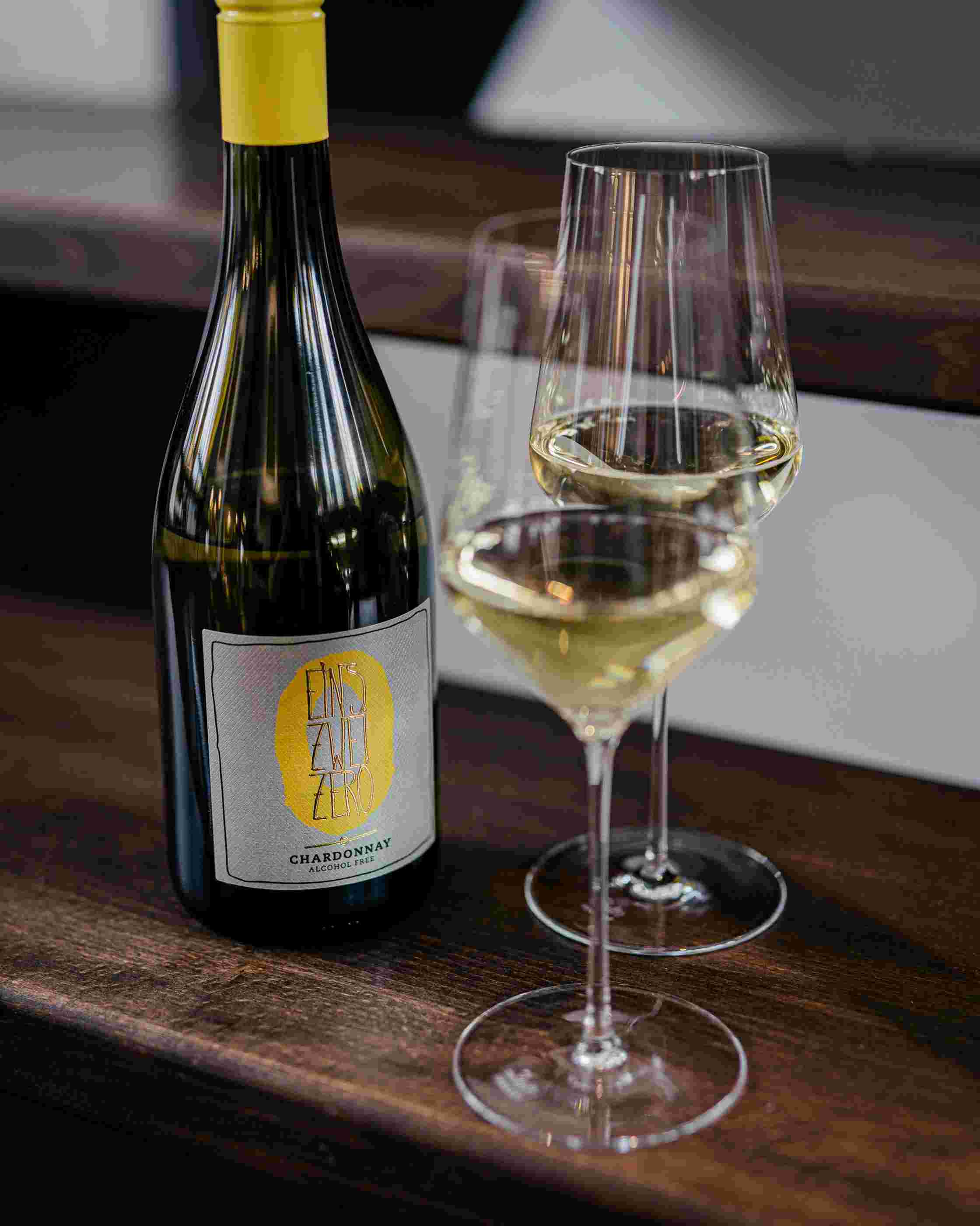 Sfeerfoto van een fles Leitz Eins-Zwei-Zero Chardonnay. Rechts staan twee wijnglazen gevuld met de Chardonnay. 