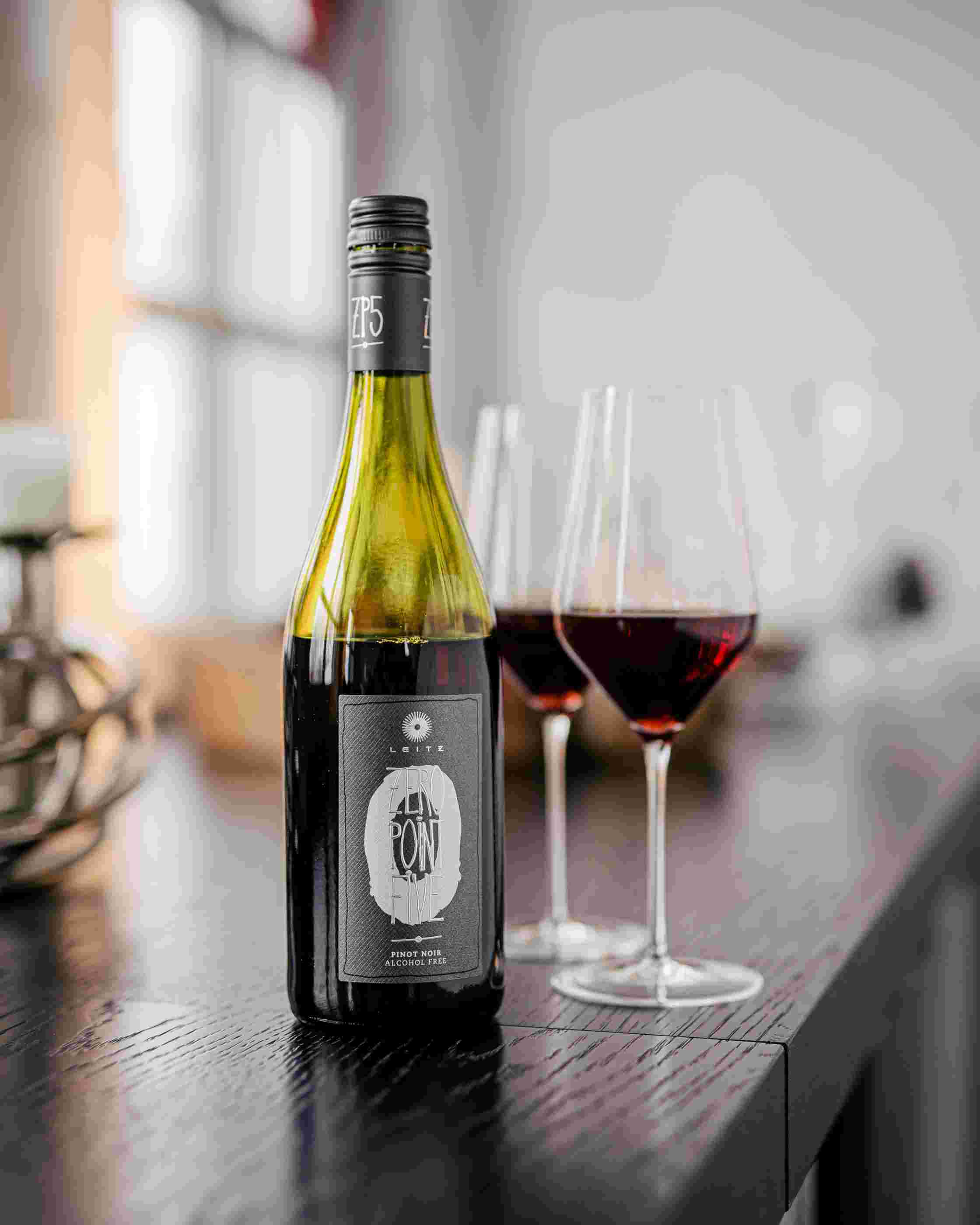 Sfeerfoto Leitz Zero-Point-Five Pinot Noir. Op een zwarte, houten tafel staat een fles Leitz Zero-Point-Five Pinot Noir. Daarachter staan twee gevulde wijnglazen.