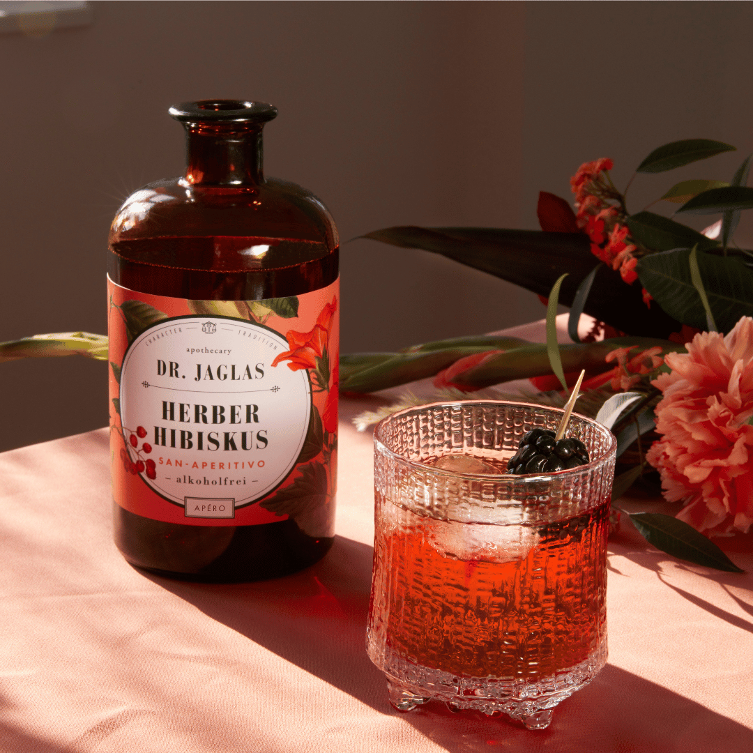 Links staat een fles Dr. Jaglas Herber Hibiscus - San Aperitivo. Rechts staat een mooi glas, waarin de Herber Hibiskus van Dr. Jaglas is ingeschonken. 