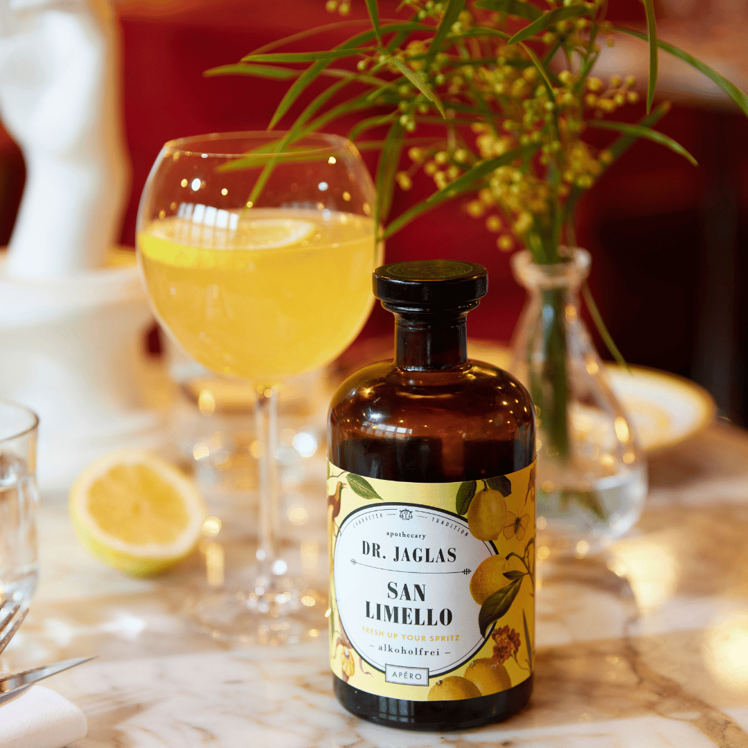 In het midden van de foto staat een fles Dr. Jaglas San Limello - Alcoholvrije Limoncello. Daarachter staat een mooi glas met alcoholvrije limoncello, gemaakt van de Dr.Jaglas San Limello.