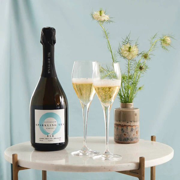 Sfeerfoto van een fles Sparkling Tea - BLÅ 0.0%. De fles staat links op een rond tafeltje. Rechts staan twee mooie glazen.