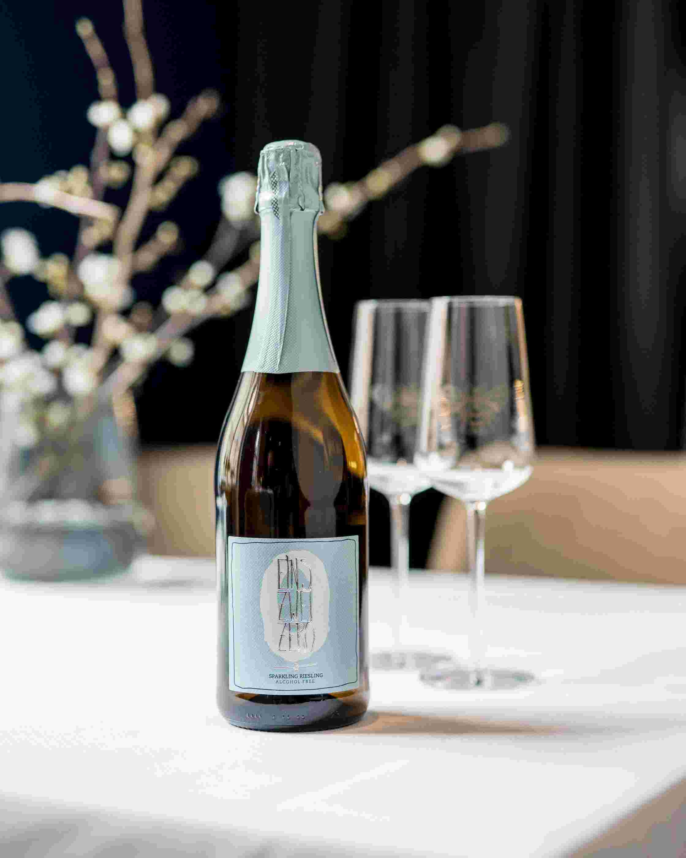 Sfeerfoto van een fles Leitz Eins-Zwei-Zero Riesling Sparkling. Op de achtergrond staan twee lege wijnglazen.