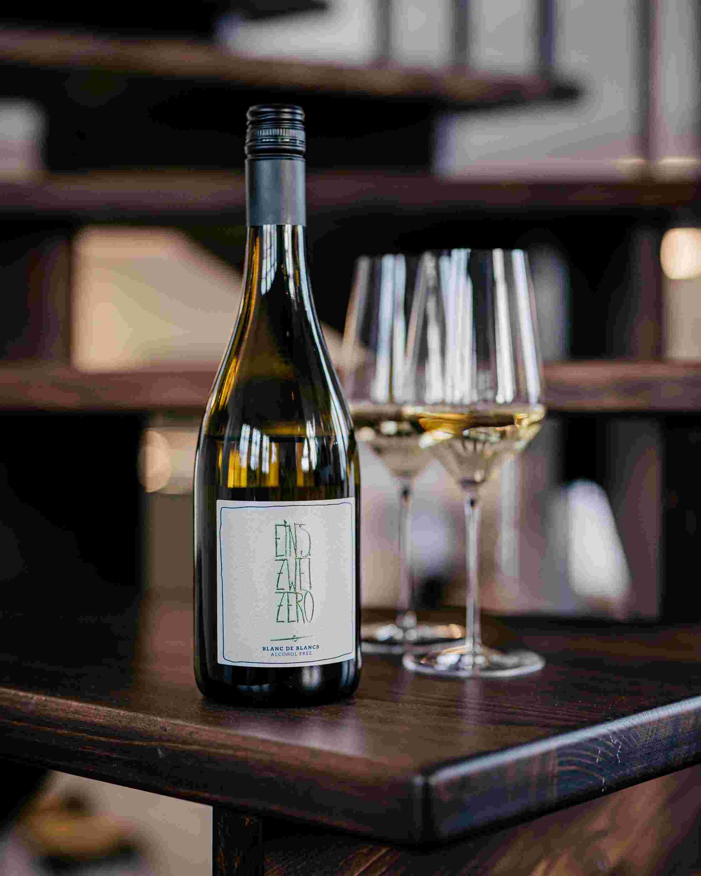 Sfeerfoto met voorop een fles Leitz Eins-Zwei-Zero Blanc de Blancs, achteraan staan twee gevulde wijnglazen op een donkere, houten tafel. 