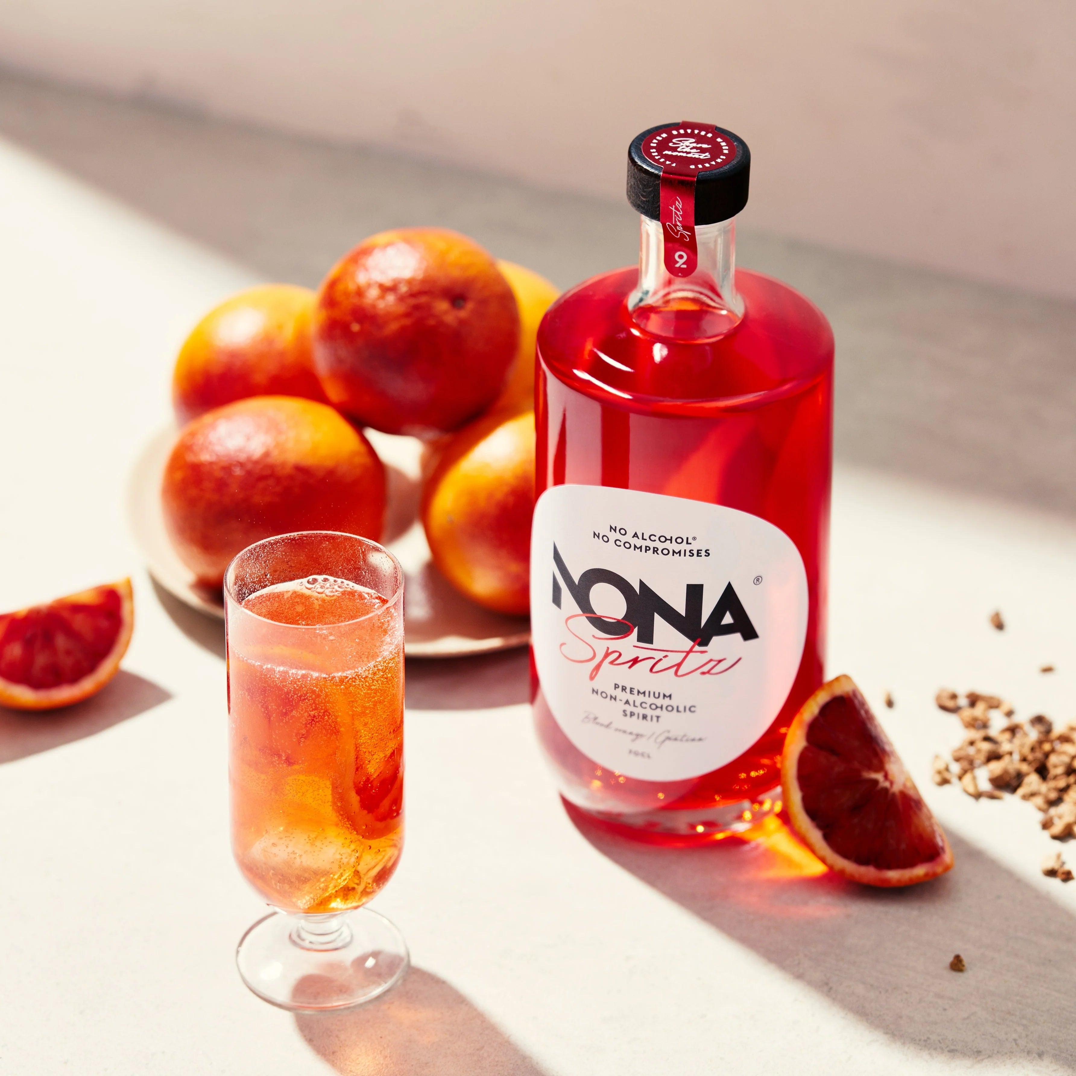 Sfeerfoto van een fles NONA Spritz 70cl. Links staat een cocktailglas gevuld met een drankje gemaakt van de Nona Spritz. Links ligt een stapel grapefruits en rechts een doorgesneden partje grapefruit. 